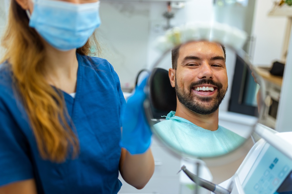 All-On-X Implants in Irvine: Patient Reviewing His Smile in the Mirror Dental patient smiling at his reflection in a hand mirror held by a team member