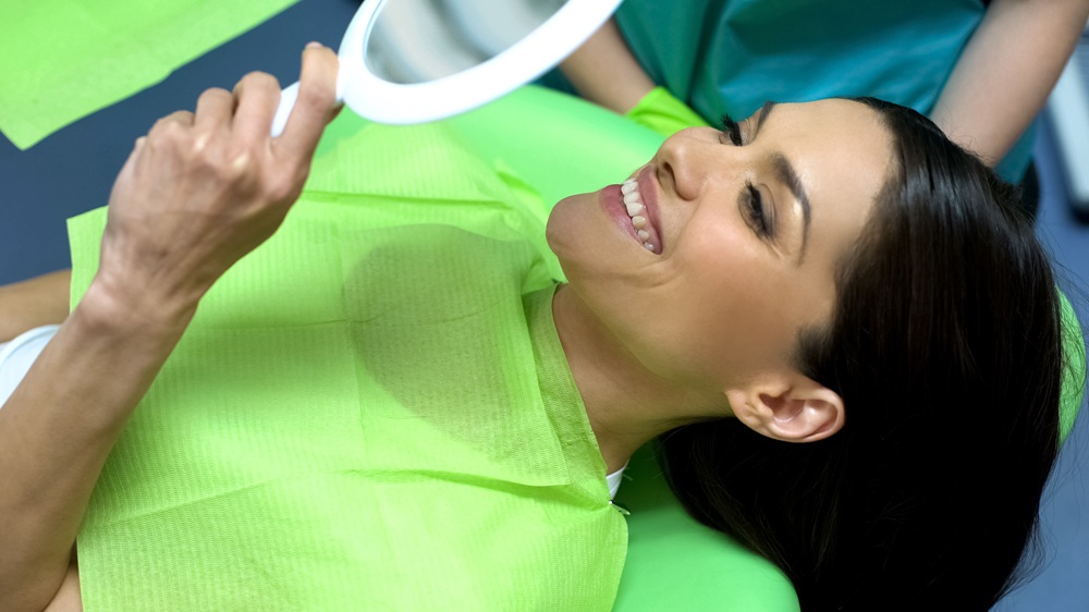 Dental patient smiling at her reflection in a handheld mirror while seated in the treatment chair